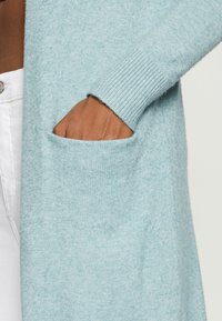 Jasnoniebieski kardigan wykonany z miękkiego materiału dziewiarskiego, z prążkowanym mankietem i boczną kieszenią po lewej stronie. Gładka struktura z delikatnym połyskiem.