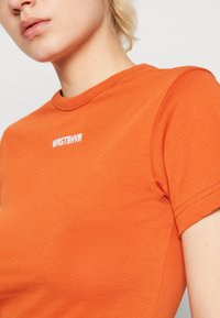 Krátký rukáv oranžového trička vyrobeného z bavlny, s kulatým výstřihem a bílým textem "WRSTBHV" na přední straně, s hladkou texturou.