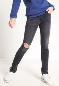 Jeans negros ajustados con rodillas desgastadas, tela ligeramente descolorida y un ajuste entallado, combinados con zapatillas blancas y una sudadera azul.