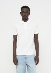 Λευκό πόλο μπλουζάκι με απαλή βαμβακερή υφή, με εφαρμοστή γραμμή, κοντά μανίκια και μικρή ετικέτα στο στήθος, συνδυασμένο με ανοιχτόχρωμο τζιν.