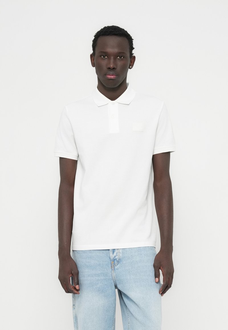 Λευκό πόλο μπλουζάκι με απαλή βαμβακερή υφή, με εφαρμοστή γραμμή, κοντά μανίκια και μικρή ετικέτα στο στήθος, συνδυασμένο με ανοιχτόχρωμο τζιν.