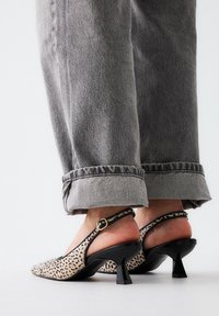 Persona vestida con jeans grises arremangados y tacones de leopardo con bloques negros, de pie sobre una superficie clara.