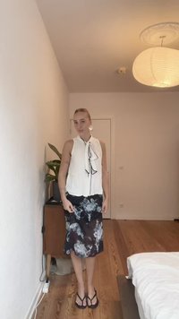 Blusa bianca senza maniche con dettaglio a fiocco, abbinata a una gonna nera floreale. Il pavimento è in legno e c'è una pianta sullo sfondo.