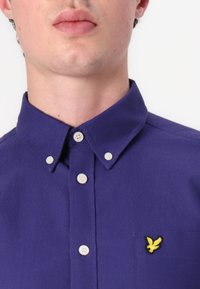 Camisa de botón morada con cuello, que presenta botones blancos y un logo de pájaro amarillo bordado en el bolsillo. Textura suave.