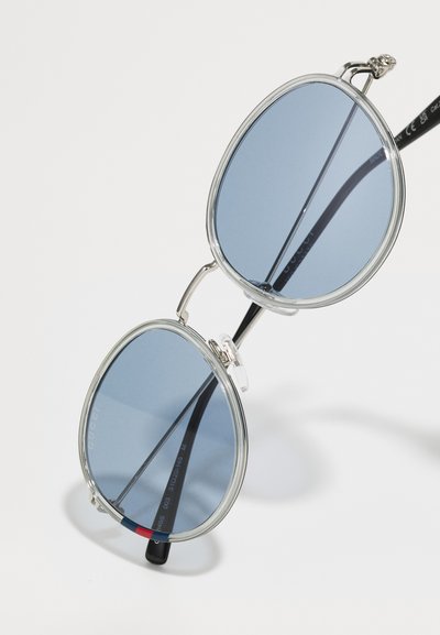 Ochelari de soare rotunzi cu lentile de culoare albastru deschis, ramă din plastic transparent și brațe negre cu accente roșii și bleumarin. Elemente metalice pentru suport.