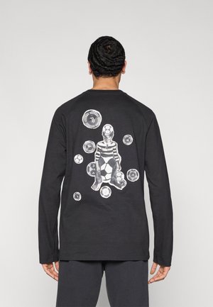Schwarzes Langarmshirt mit einem grafischen Design auf dem Rücken, das eine Figur mit einem gestreiften Muster und weißen sowie grauen Fußbällen zeigt.