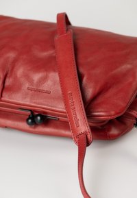 Rotte Ledertasche mit glatter Textur, ausgestattet mit einem langen Trageriemen und schwarzen Hardware-Details. Zu den Merkmalen gehören eine Markenprägung.