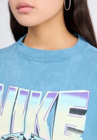 Mėlynas medvilnės džemperis su ryškiu „NIKE“ tekstu gradientinėmis spalvomis. Dizainas apima apvalų kaklą ir minkštą, tekstūruotą paviršių.