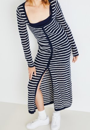 Kvinde iført en lang kjole med knapper i navy og hvide striber med en sidespalte, hvide sneakers og hvide sokker, stående mod en ensfarvet væg.