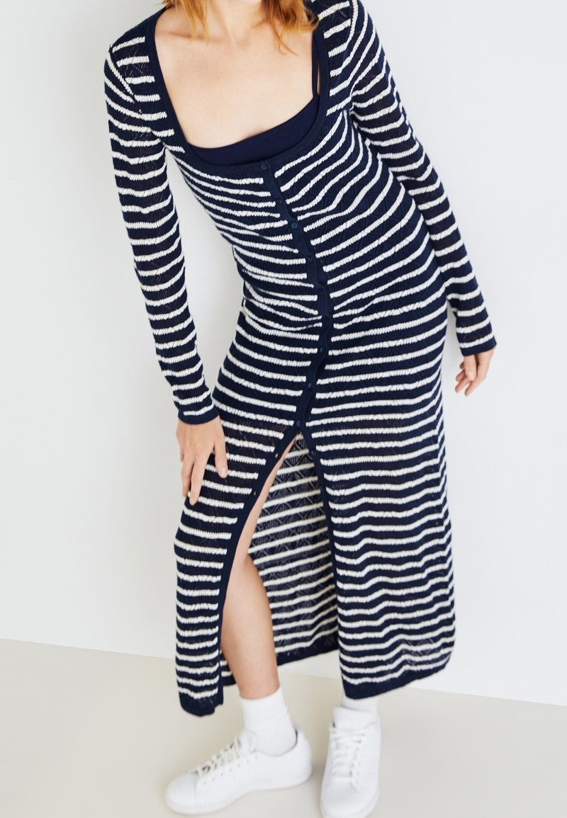Femme portant une longue robe boutonnée rayée bleu marine et blanche avec une fente sur le côté, baskets blanches et chaussettes blanches, debout contre un mur uni.