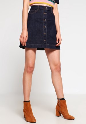 Kvinde iført mørk denimnederdel med knapper foran, stribet top og brune ruskindsstøvler til anklen, stående mod en ensfarvet lys baggrund.