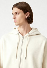 Felpa con cappuccio color crema realizzata in tessuto morbido, con zip frontale completa, cappuccio regolabile con cordino e dettagli in metallo argentato.