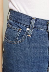 Jeans de ganga de cintura alta em azul escuro com costura laranja. As características incluem um bolso frontal, botão de metal e tecido texturizado.