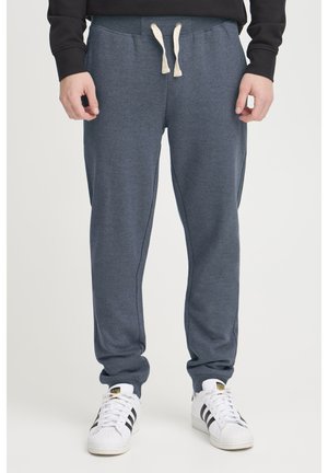 Pantalones de chándal azul marino fabricados con un tejido suave, con cintura de cordón, acentos en las costuras laterales y un diseño de pierna ajustada. Combinados con zapatillas blancas.