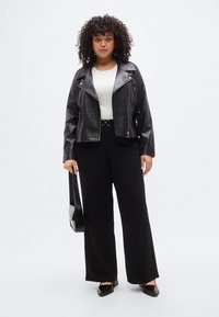 Μαύρο μπουφάν μοτοσικλετιστή από δερματίνη, λευκό τοπ, μαύρο παντελόνι με φαρδιά μπατζάκια, μαύρα μυτερά παπούτσια και μαύρη τσάντα χειρός. Σγουρά μαλλιά, ουδέτερο φόντο.