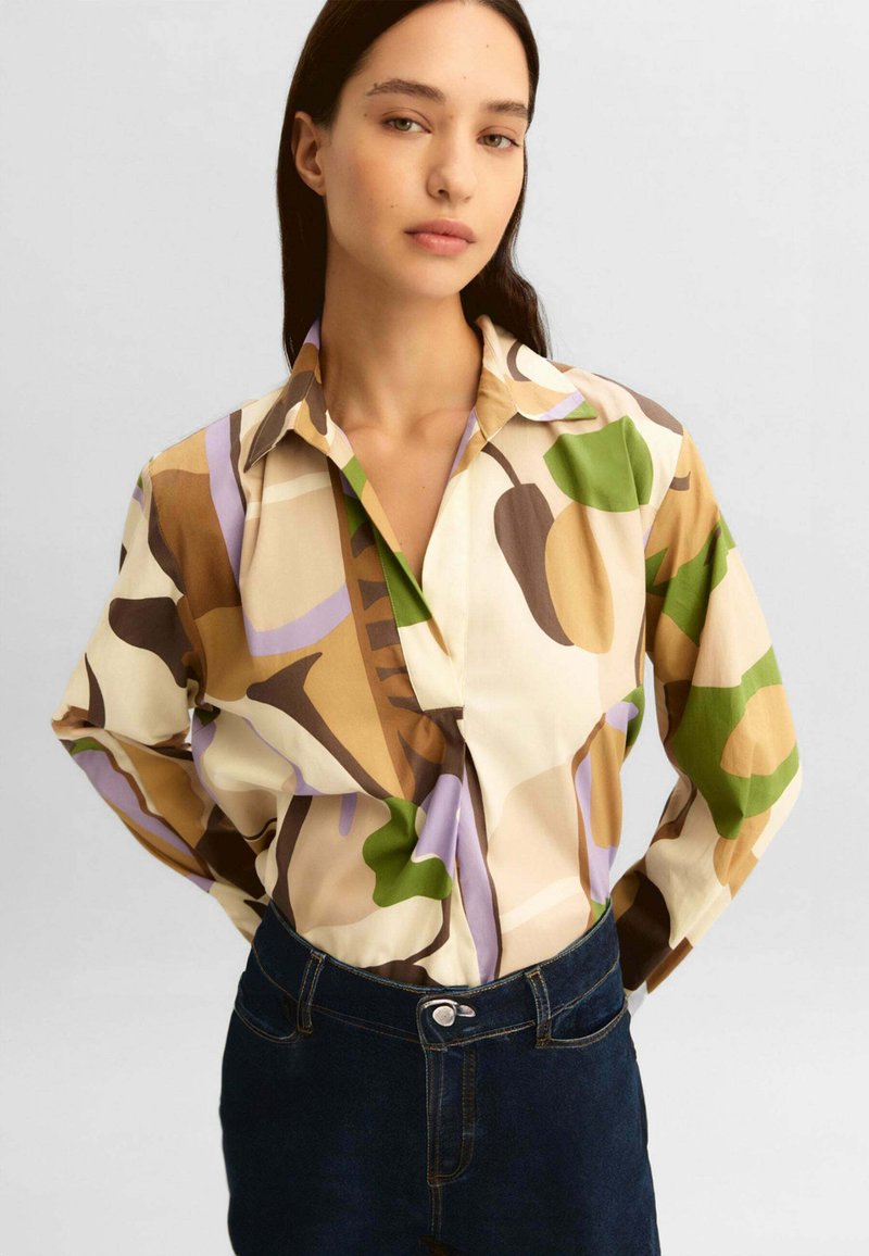 Donna con capelli lunghi e scuri che indossa una camicetta con motivo astratto multicolore e jeans blu scuro a vita alta, in piedi con le mani dietro la schiena.