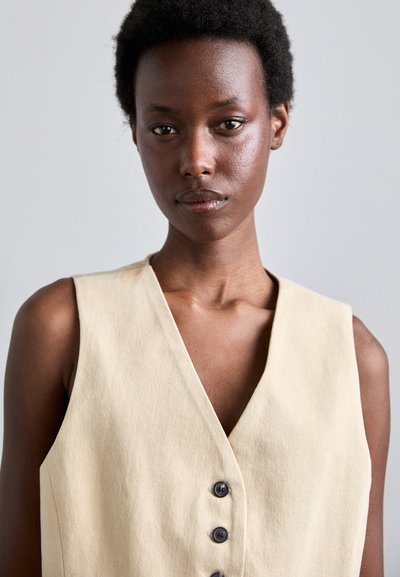 Beige mouwloze vest met een diepe V-hals, voorzien van vier donkere knopen, gemaakt van gestructureerde stof, en biedt een getailleerde silhouet.