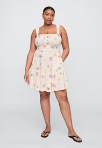 Vestido bege claro com alças ajustáveis, cintura franzida e saia em camadas com padrões coloridos de conchas e sol. Usado com chinelos pretos.