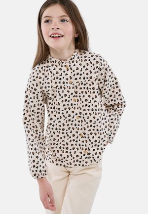 Jeune fille aux longs cheveux bruns portant une chemise crème à boutons imprimée léopard et un pantalon beige clair, souriante et regardant légèrement sur le côté.