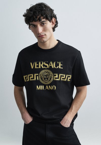 Sort bomulds-T-shirt med guldfarvet tryk af teksten "VERSACE MILANO" og et centralt Medusa-logo, med et græsk nøglemønster under logoet.