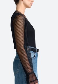 Zwarte top met doorschijnende, gemusterde lange mouwen en een slim fit. Gecombineerd met hoog-gewaste blauwe denim jeans en een zwarte riem.