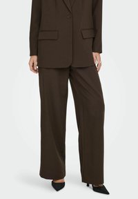 Dunkelbraune, maßgeschneiderte Hose mit weitem Bein, ausgestattet mit einem sauberen Finish und glatter Textur, kombiniert mit spitzen schwarzen Absätzen.