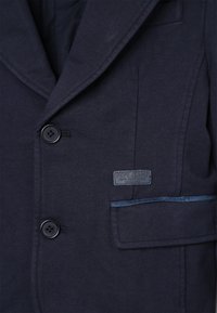 Blazer bleu marine en tissu texturé, doté de deux boutons, d'une poche poitrine et d'une étiquette Pepe Jeans avec une couture d'accent sur la poche.