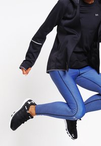 Zwarte capuchonjack met reflecterende accenten, zwart shirt, blauwe gestructureerde leggings en zwarte sportschoenen met witte zolen, in een springpose.