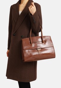 Borsa con motivo coccodrillo marrone, forma strutturata, due manici, tasca frontale piatta con un accento metallico, tenuta da una persona in un cappotto marrone scuro.