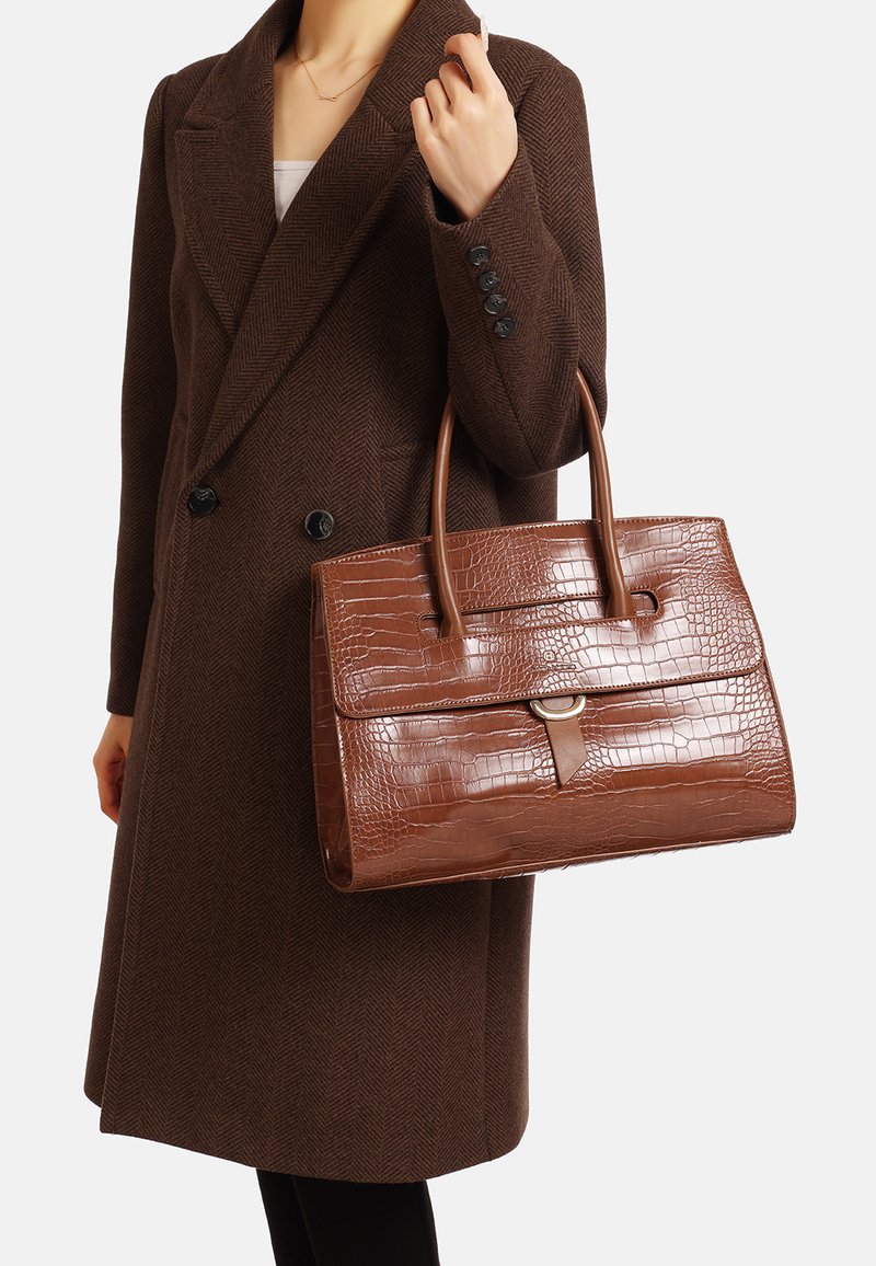 Borsa con motivo coccodrillo marrone, forma strutturata, due manici, tasca frontale piatta con un accento metallico, tenuta da una persona in un cappotto marrone scuro.