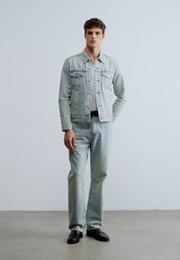 Ljusblå jeansjacka med två bröstfickor, vit t-shirt under och ljusblå jeans, kombinerat med svarta skor. Enkel design.