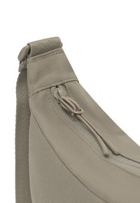 Gros plan sur un coin de sac en tissu gris avec fermeture éclair et détail de corde de tirage, accompagné d'une boucle de sangle réglable.