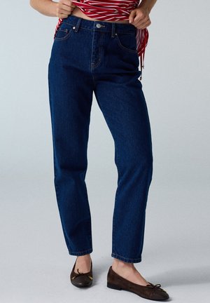 Persona che indossa jeans blu scuro a gamba dritta, ballerine marroni scure e una maglia a righe rosse e bianche sollevando leggermente l'orlo.