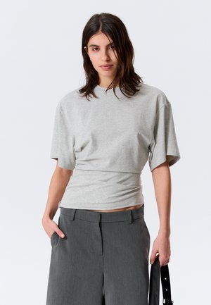 Kvinne med mørkt hår iført en lysegrå tettsittende t-skjorte og mørkegrå bukser, holder en svart stropp i høyre hånd.