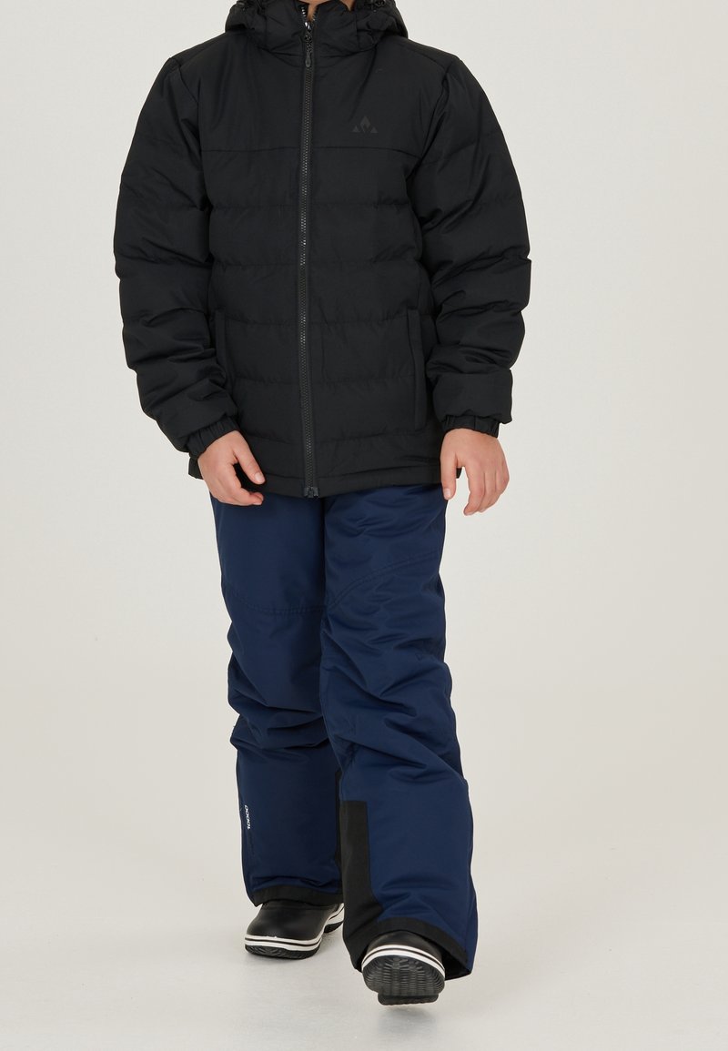 Enfant portant une doudoune noire à capuche, un pantalon de neige bleu marine et des bottes d'hiver noires marchant vers l'avant sur un fond clair uni.