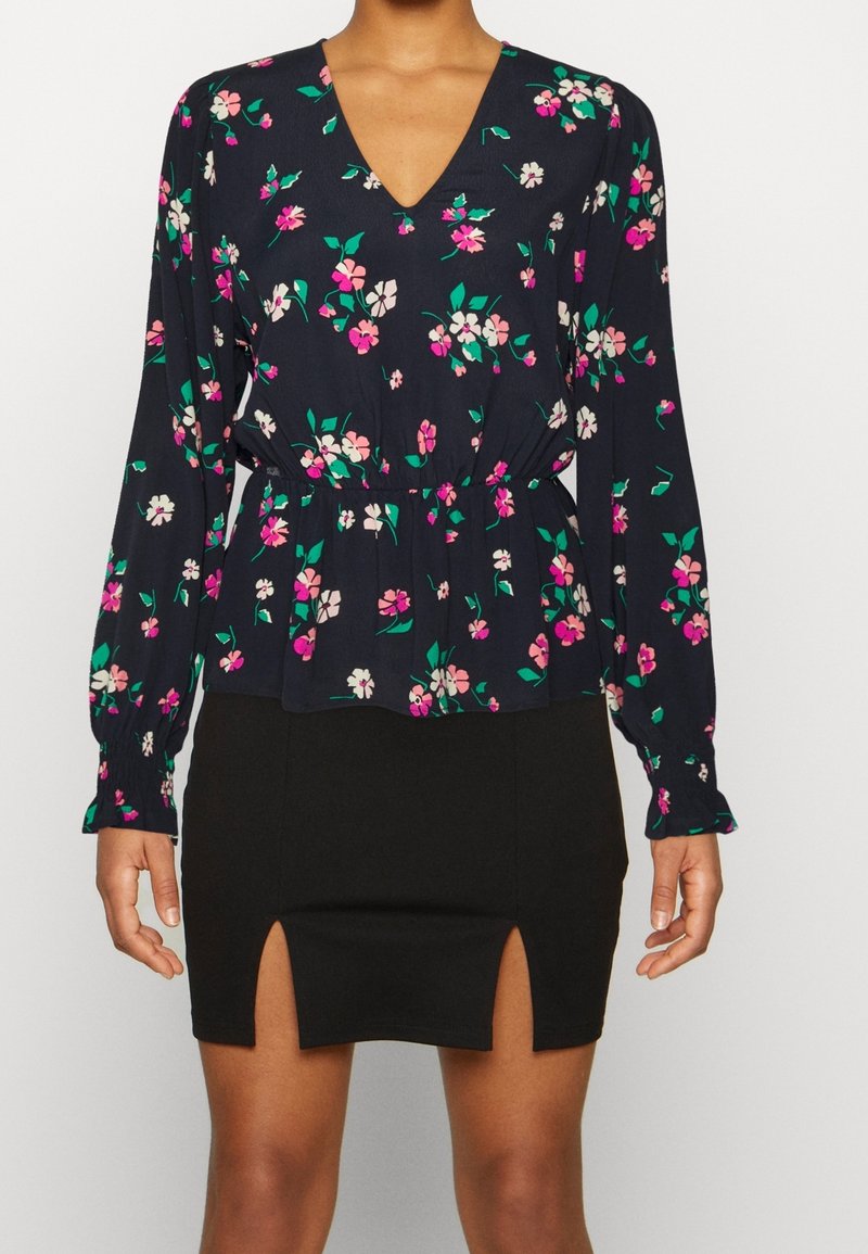 Kukallinen pusero tummansinisestä kankaasta, jossa on pitkät hihat, V-pääntie ja kerroksittain laskostettu vyötärö. Yhdistetty mustan hameen kanssa, jossa on sivuleikkaukset.