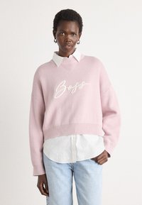 Rosa strikket genser med "Boss" brodert i hvitt, med v-hals, senket skuldre og ribbestrikket kant, lagd over en hvit skjorte.