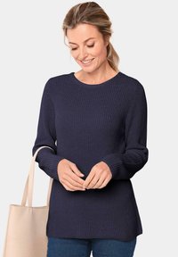 Marineblauer Strickpullover mit Rundhalsausschnitt, Rippbündchen und Nahtdetails, kombiniert mit einer hellbeigen Tragetasche.