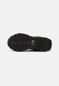 Black rubber hiking shoe outsole with deep treads for traction. Features a textured surface and a yellow accent among the tread patterns.