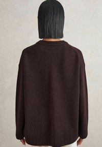 Une personne avec des cheveux noirs lisses jusqu'aux épaules porte un pull en tricot à manches longues de couleur marron foncé, de dos contre un fond uni.