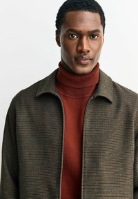 Braune Hahnentritt-Jacke mit Reißverschluss, getragen über einem roten, gerippten Rollkragenpullover. Glatte Stoffstruktur und mit Kragen.