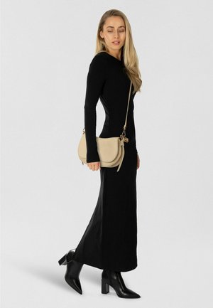 Robe noire à manches longues avec une silhouette ajustée, associée à un sac à main en cuir beige clair. Le modèle porte des bottines noires à talons.