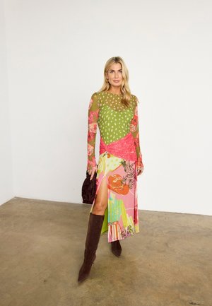 Kvinde med langt blondt hår iført en farverig mønstret kjole med et lårslids, brune knæhøje støvler og med en mørkebrun håndtaske.