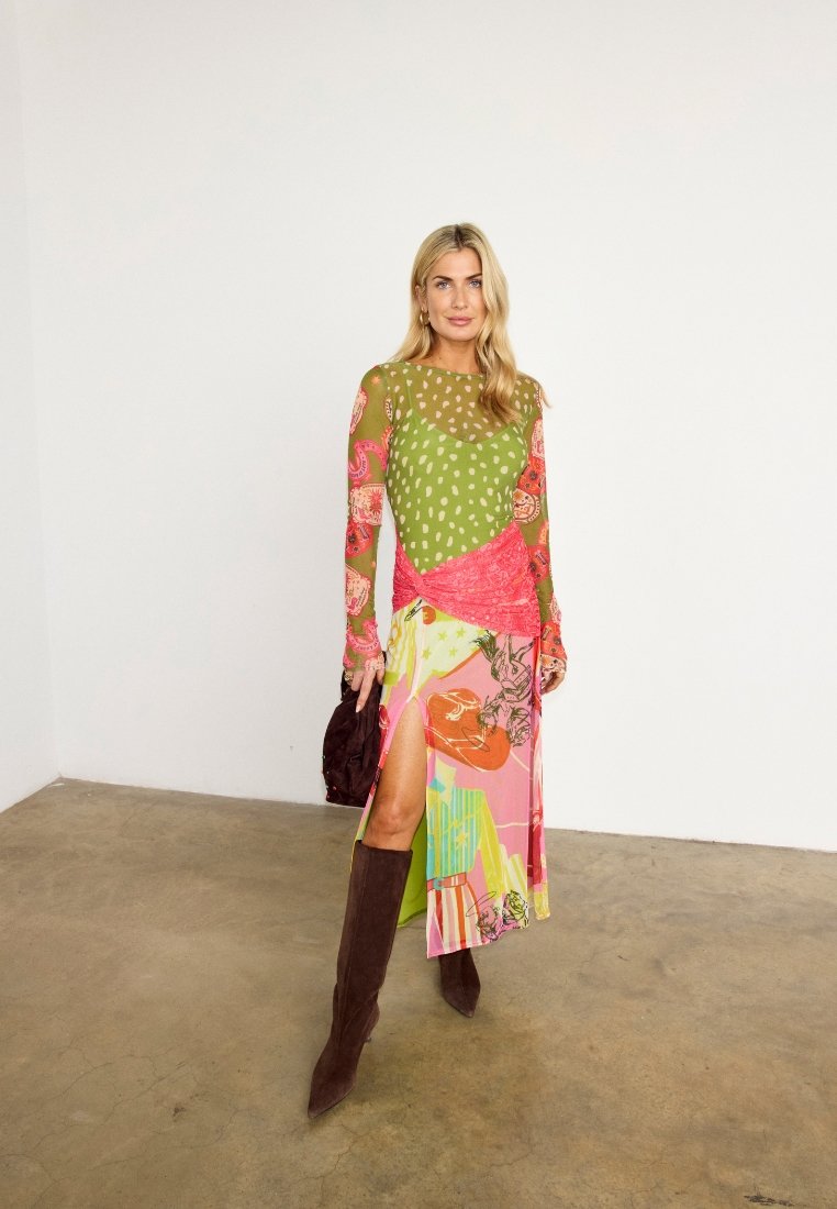 Femme aux longs cheveux blonds portant une robe colorée à motifs avec une fente sur la cuisse, des bottes marron jusqu'aux genoux et tenant un sac à main marron foncé.