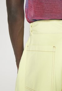 Brazo izquierdo de una persona que lleva una camiseta de malla rosa y morada metida en pantalones amarillos pálidos con un bolsillo trasero visible.