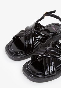 Sandali in pelle verniciata nera con cinghie incrociate, suola piatta e dettagli tondeggianti. Il design presenta una finitura lucida e una silhouette elegante.
