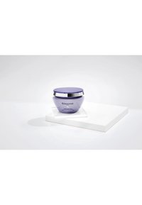Lavender Kérastase Haarmasken-Glas mit silbernem Deckel auf weißen geometrischen Plattformen vor einem schlichten hellen Hintergrund präsentiert.
