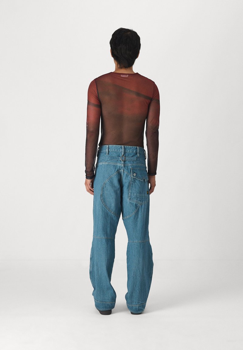 Top marrón semitransparente de manga larga con un diseño ajustado y matices rojizos en contraste, combinado con unos pantalones vaqueros de denim azul sueltos con un bordado único.