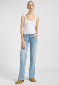 Top de tirantes blanco con canalé, vaqueros acampanados de color azul claro y zapatos slip-on de color tostado. Los vaqueros tienen un lavado ligeramente desgastado y un diseño de corte recto.