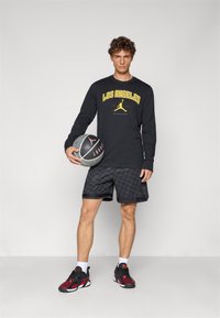 Langærmet sort skjorte med "LOS ANGELES" og logo i gult, parret med mønstrede sorte shorts og sorte sneakers med røde accenter, mens en basketball holdes.